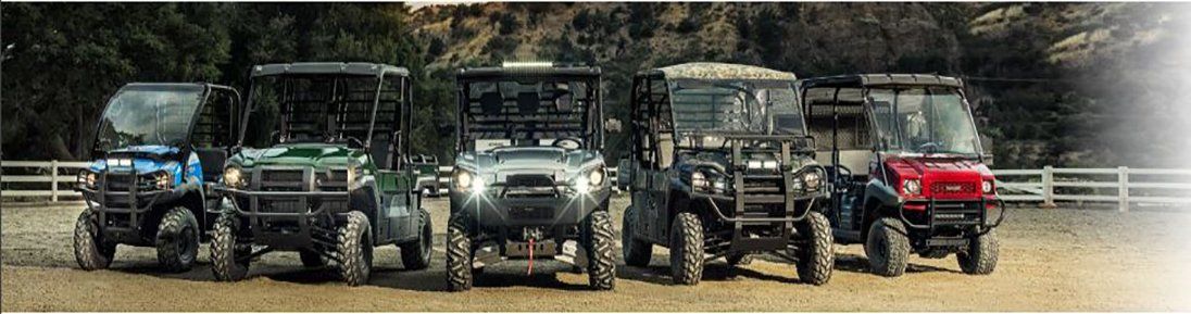 Six utility vehicles parked side-by-side on a dirt field with a fence and trees in the background.