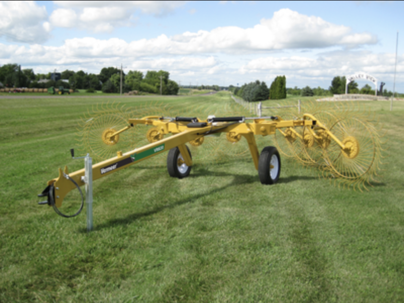 Yellow hay rake on a green grassy field under a cloudy blue sky.