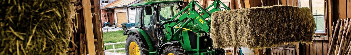 A John Deere tractor is in a barn with a hay bale and crop on the side.