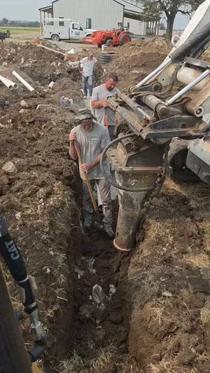 Workers guide a large drilling machine into a muddy trench at a construction site.