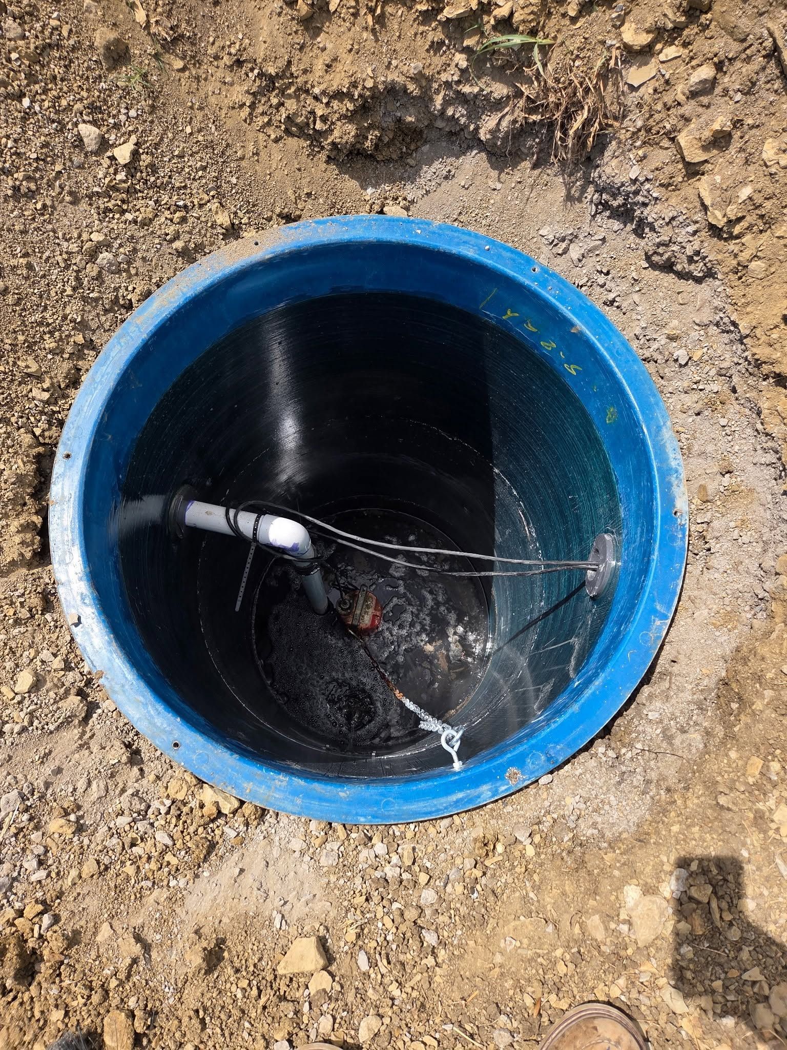 Blue-rimmed open pipe or manhole in dirt, showing dark interior with water inside.