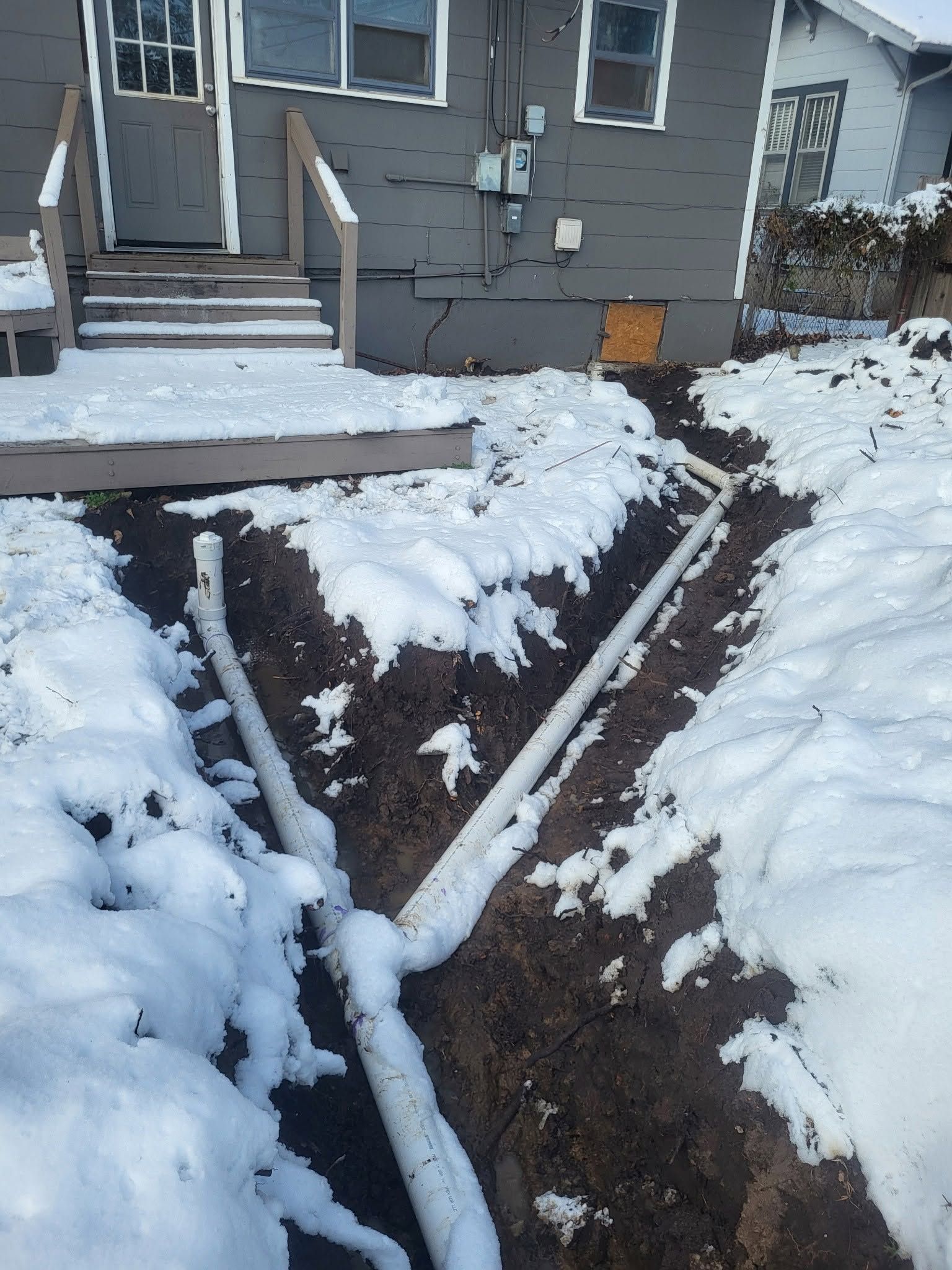 Snow-covered trench with a diagonal pipe beside a house steps and siding