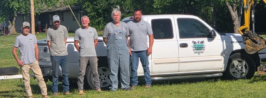 Five workers in gray shirts stand beside a white utility truck on grass in front of trees.