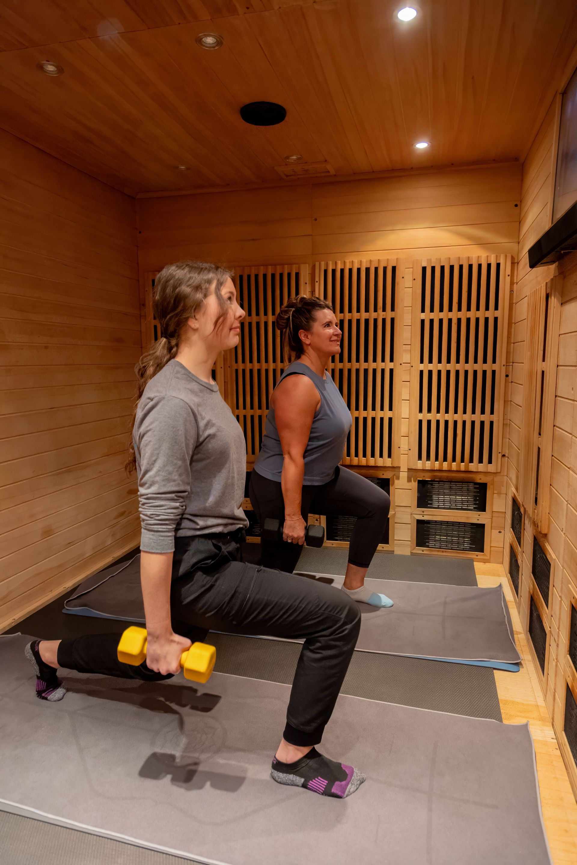 Two individuals perform lunges with dumbbells inside a wood-paneled room.