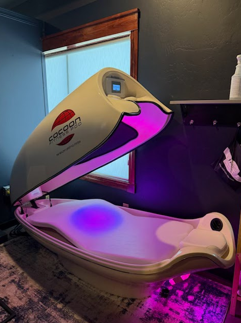 A Cocoon Fitness Pod wellness machine with an open lid, glowing purple and pink interior, in a dark, neutrally toned room.
