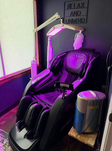 A black massage chair in a room with purple lighting, a hanging UV lamp, and a portable oxygen concentrator on a side table.
