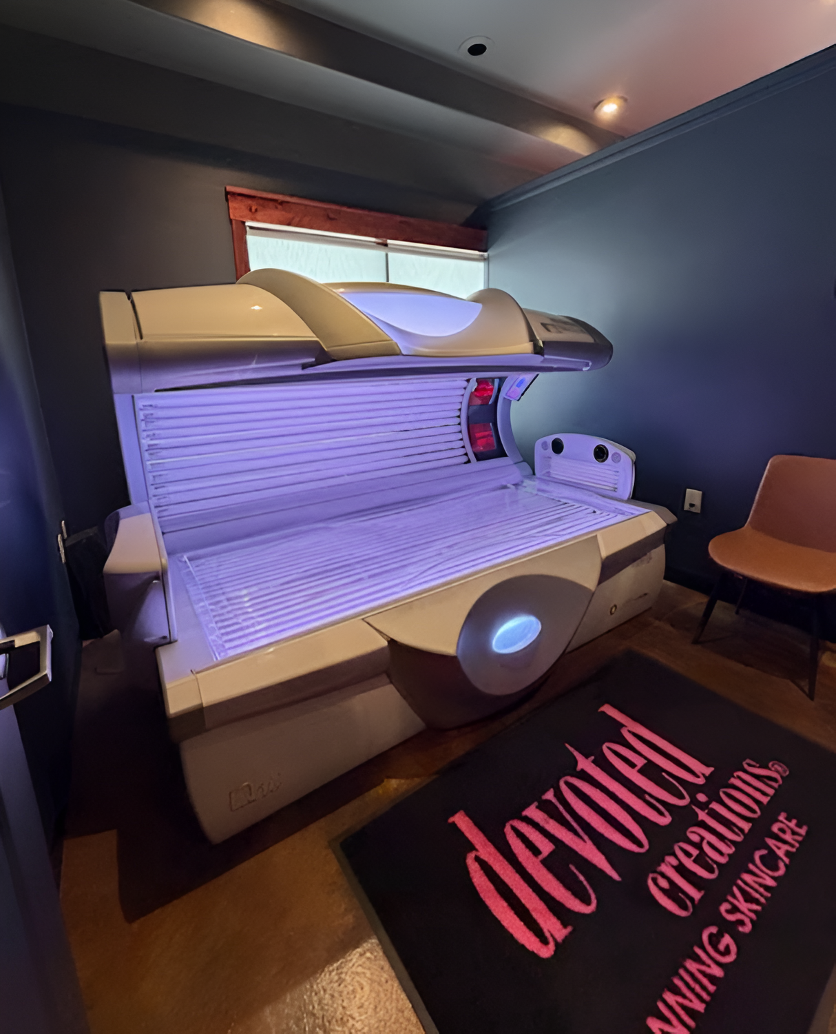 A horizontal tanning bed glows in a dark blue room next to a rug labeled