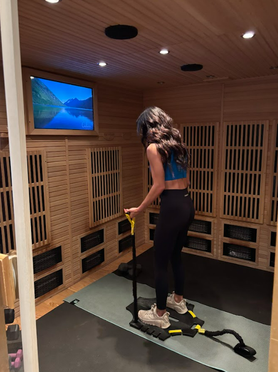 A person stands on a resistance training platform inside a wood-paneled infrared sauna, looking at a wall-mounted screen.