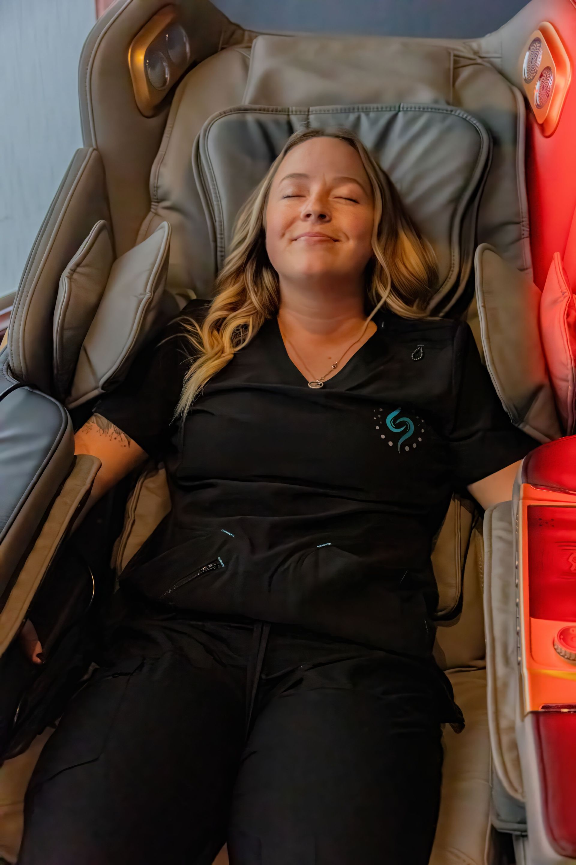 A person with long blonde hair relaxes with eyes closed in a padded massage chair while wearing a black uniform.