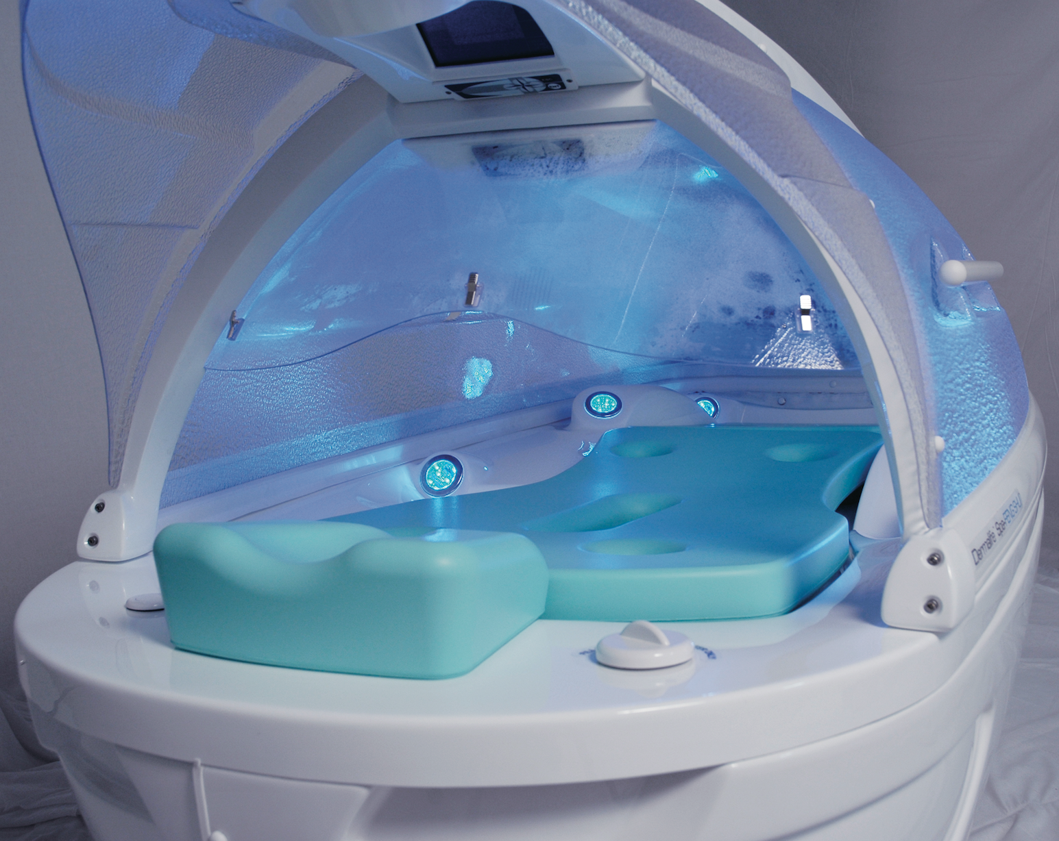 A white spa capsule with a turquoise headrest and interior, illuminated by blue light in an open, reclined position.