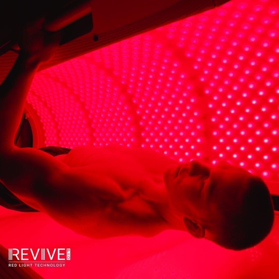 A person lies inside a tunnel illuminated by bright red LED lights, using REVIVE red light therapy technology.