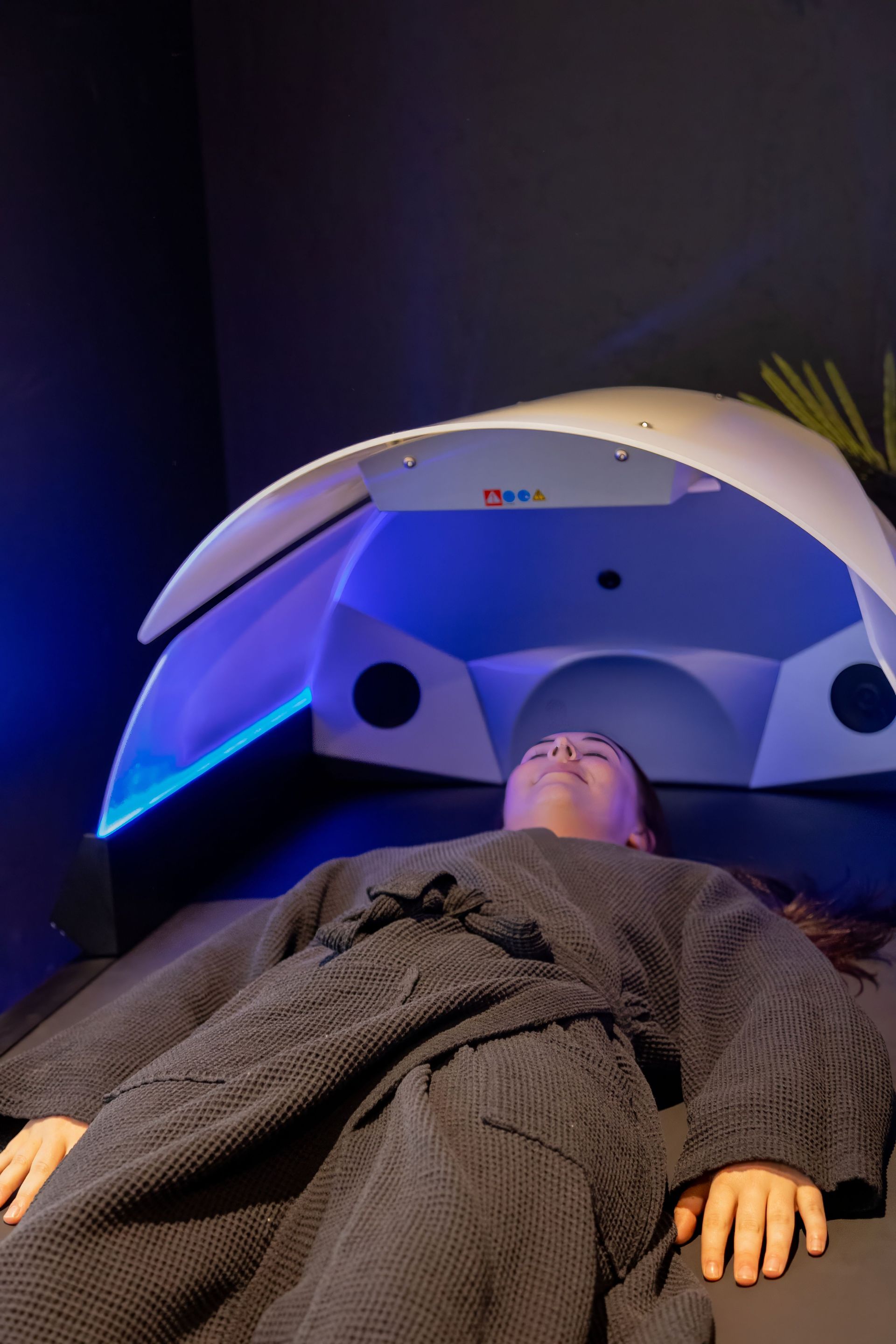 A person in a robe lies under a glowing blue LED light therapy dome in a dimly lit room.