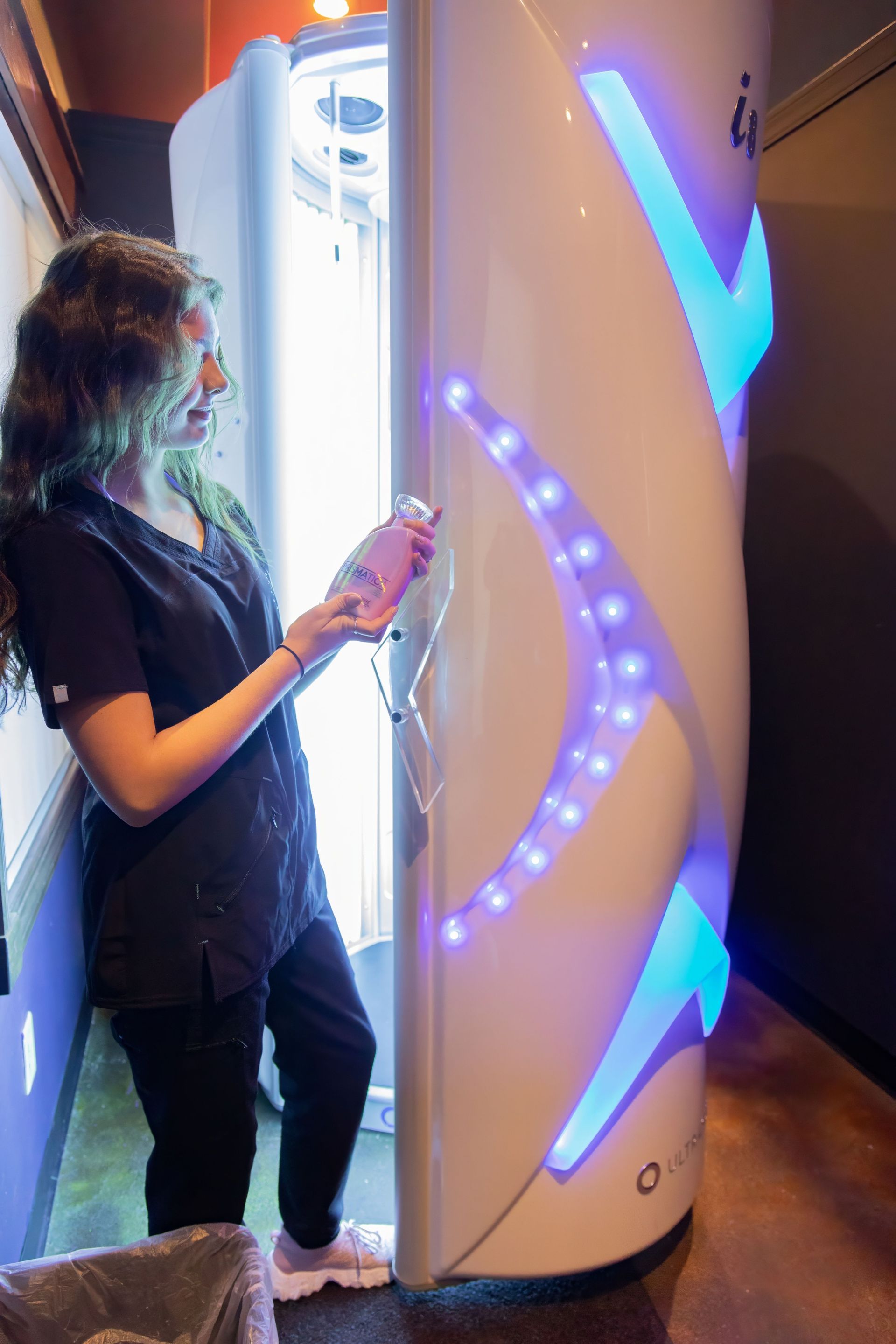 A person in dark scrubs holding a remote next to an illuminated vertical tanning booth in a salon.