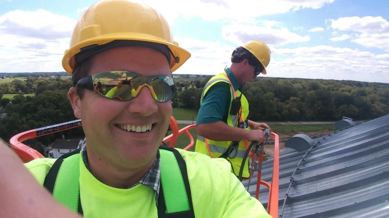 Two construction workers wearing hard hats and safety vests are working on a roof.