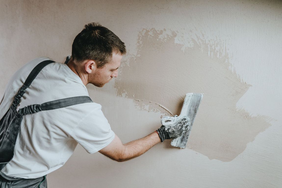 Man smoothing wet plaster on a wall with a trowel, wearing a white shirt and gray suspenders.