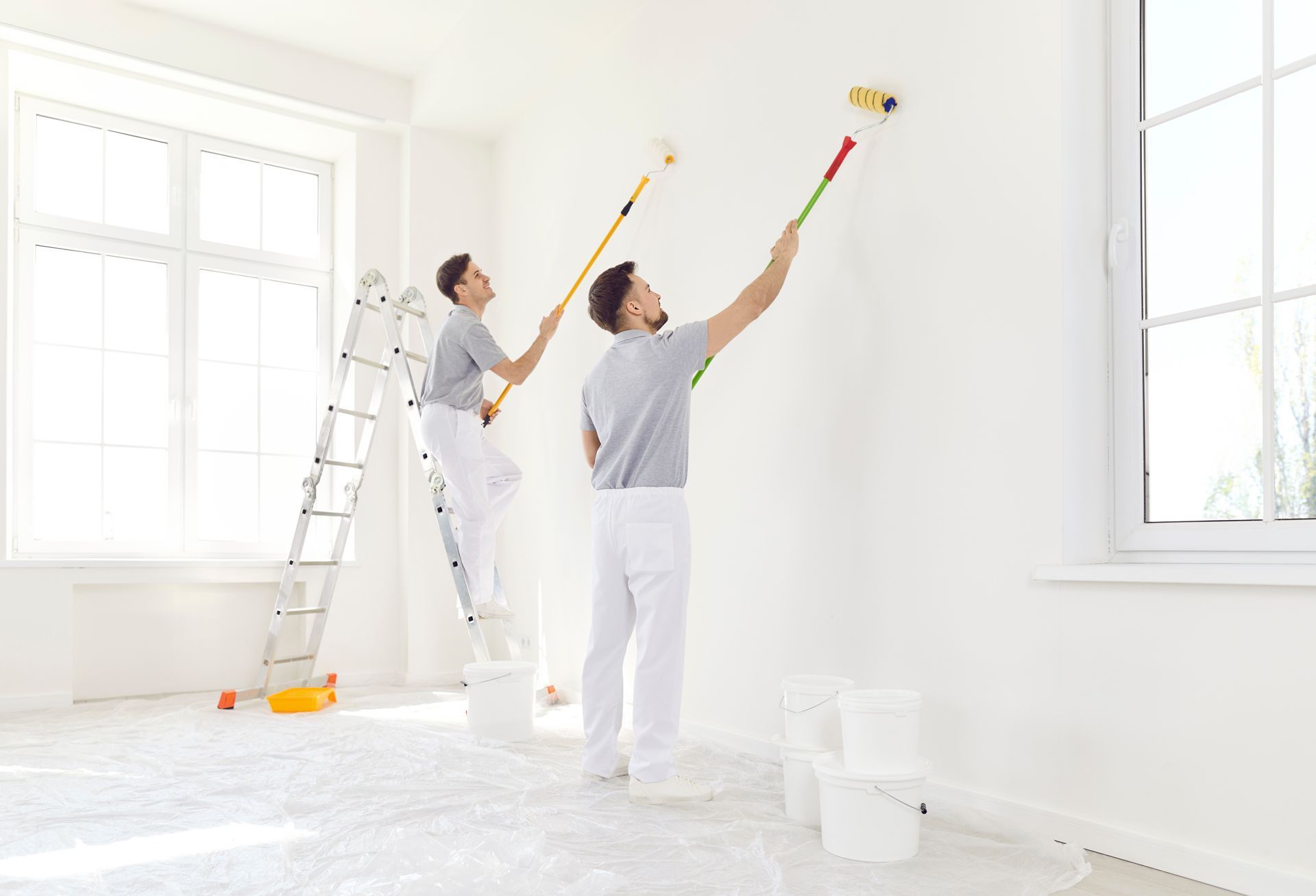Two painters in white painting a bright room, using rollers near a ladder and paint buckets.