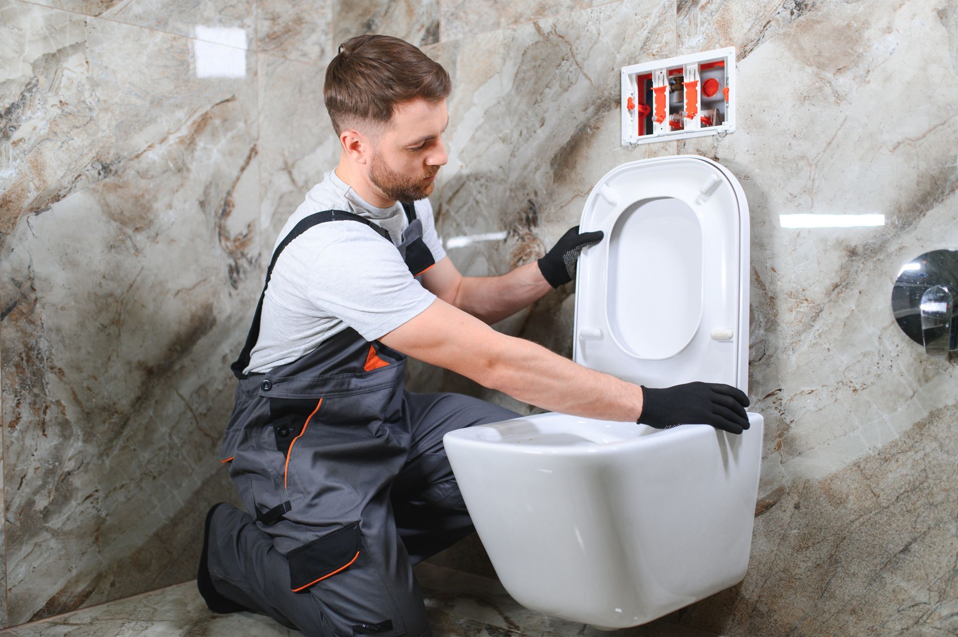Plumber in gray overalls installing a white urinal in a tiled bathroom