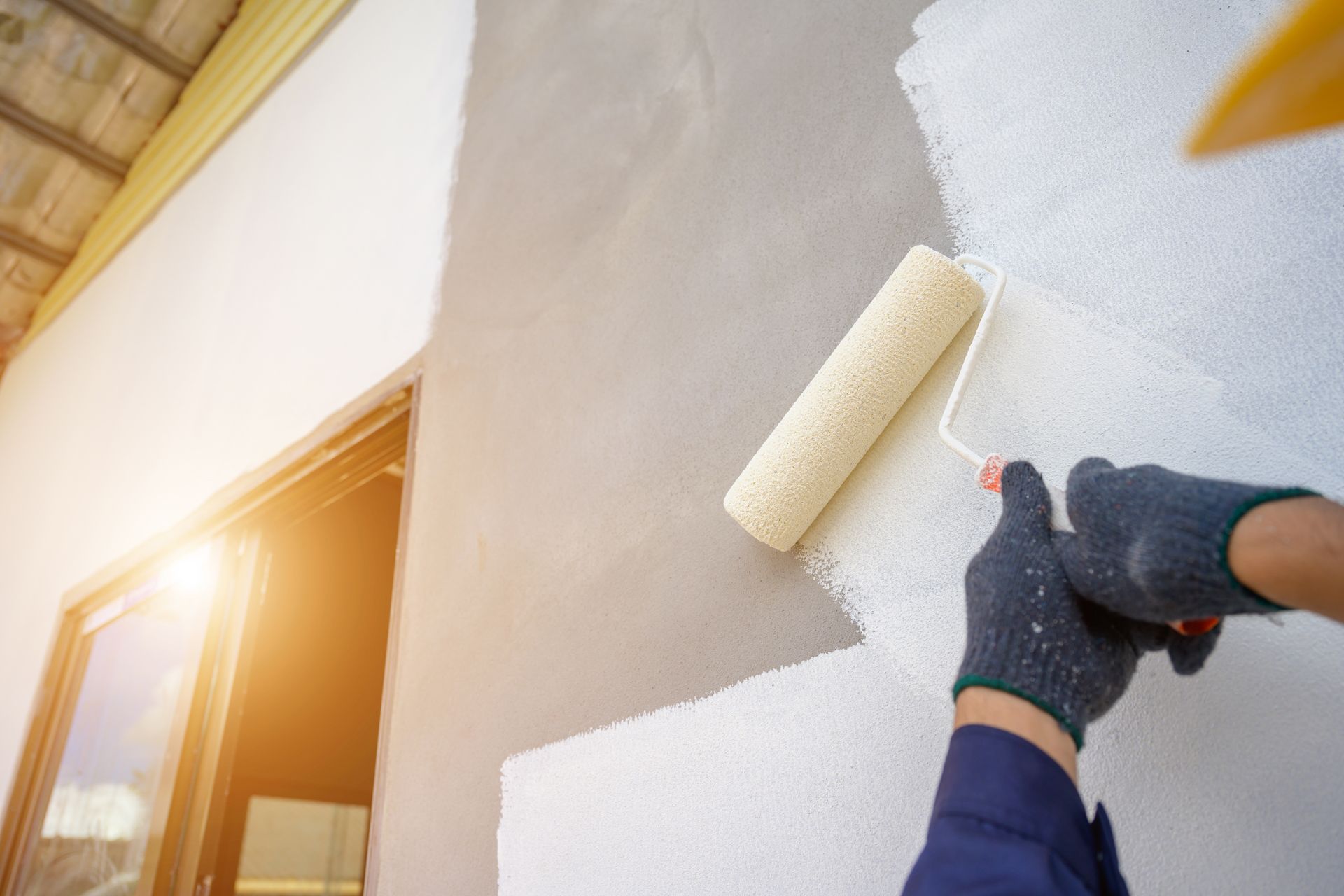 Person painting a white exterior wall with a roller near a window in bright sunlight