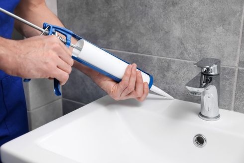 Hand using a caulking gun to apply white sealant around a bathroom sink faucet