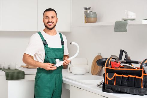 Plumber in green overalls holding a white pipe in a bright kitchen with tools on the counter