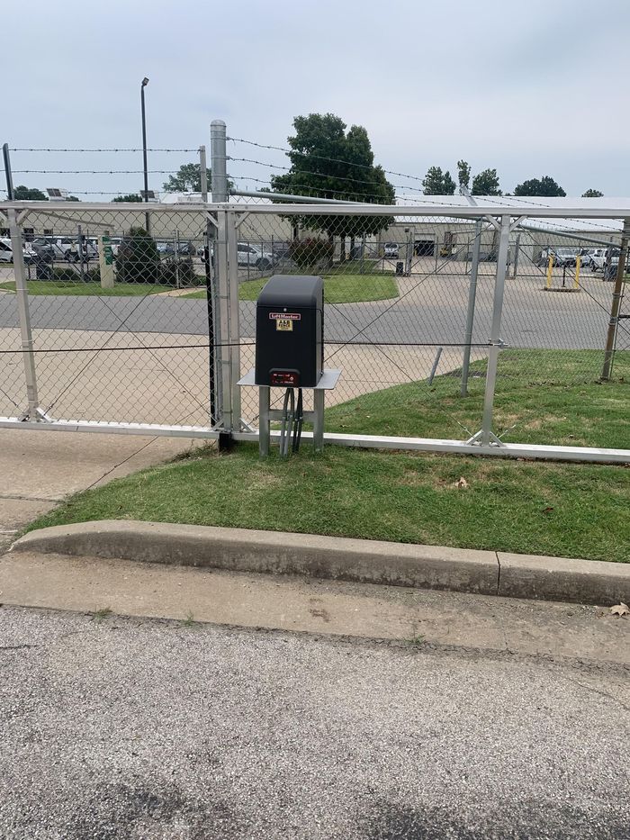 Chain-link fence with a black mailbox and trash can beside a road, with a grassy lot in the background