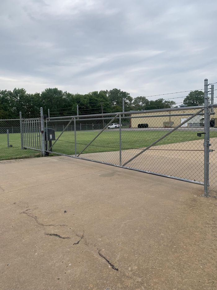Chain-link gate opening onto a paved lot near a beige building under a cloudy sky.