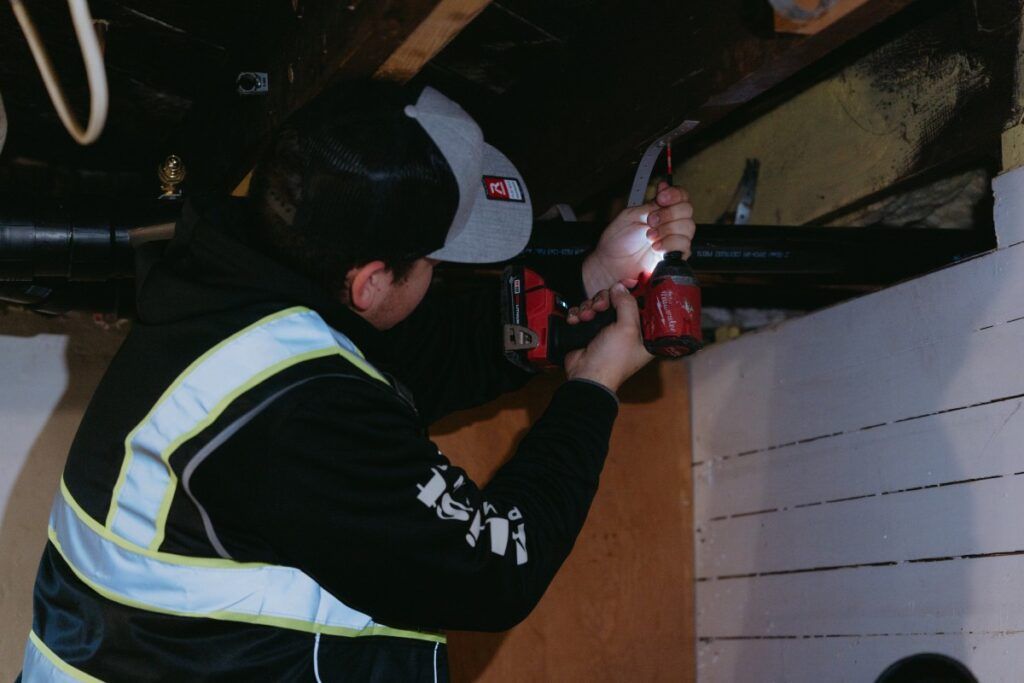 Person in reflective jacket using a power drill on a pipe in a basement.
