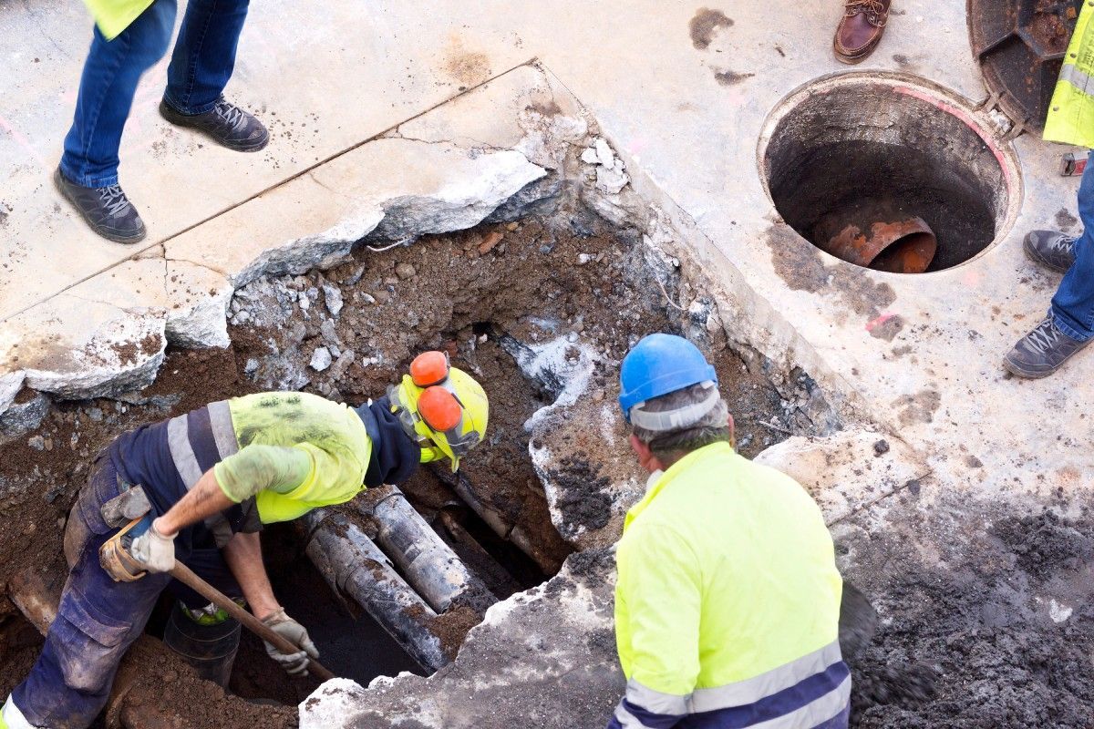 Workers in hard hats repairing a dug-up street trench near a manhole.
