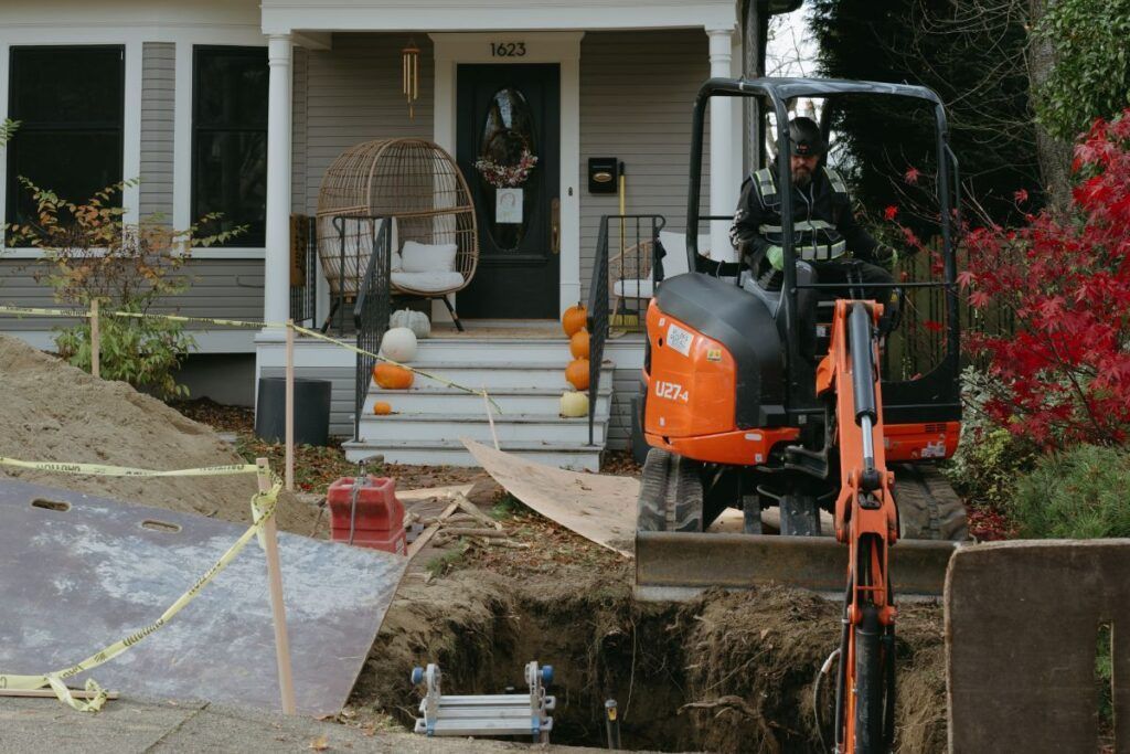 A man is driving an excavator in front of a house for plumbing.