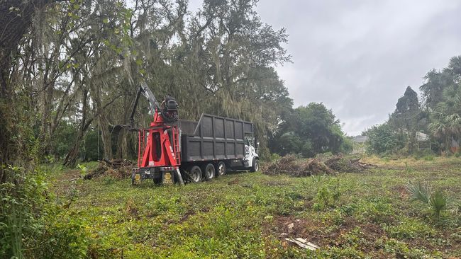 A truck with a crane loads brush in a grassy area near trees under an overcast sky.