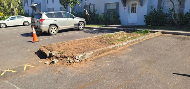 A parking lot with a broken curb, orange cone, SUV, and building in the background.