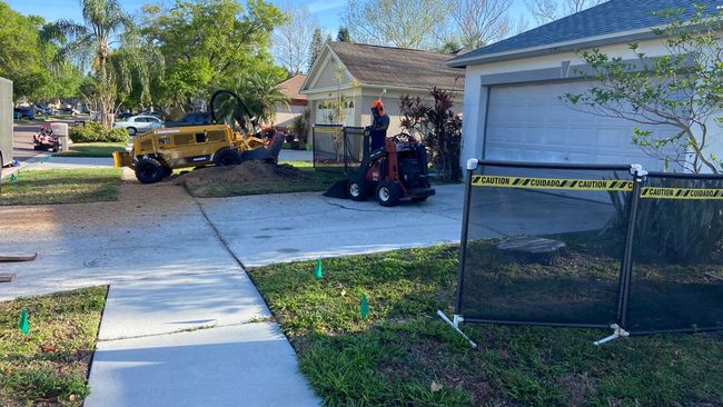 Tree stump removal: Man operates machinery on a residential driveway. Yellow barriers and safety tape surround the work area.