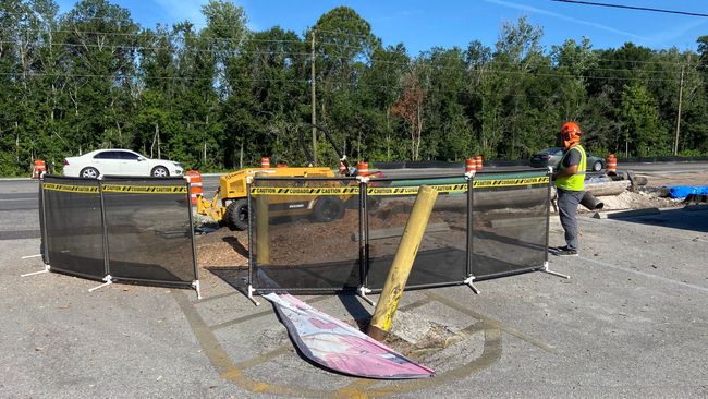 Construction site in a parking lot with a worker and vehicle behind a black safety barrier; a white car drives past.