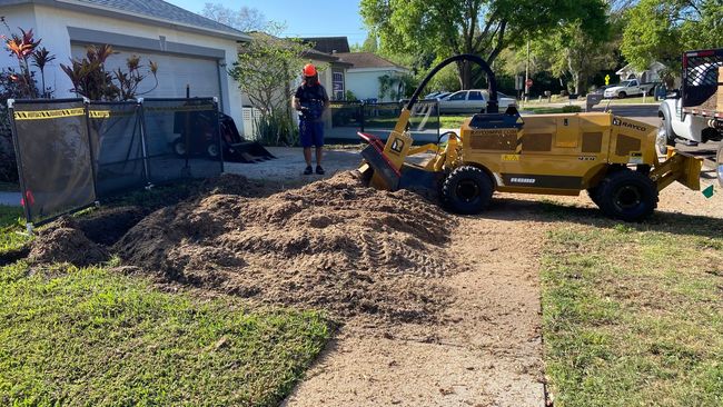 A person operates a yellow stump grinder on a driveway, creating wood chips.