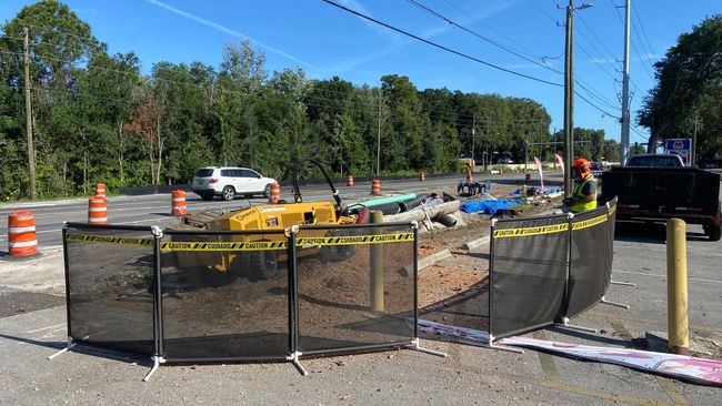 Construction site with black mesh fencing, orange cones, machinery, and workers.