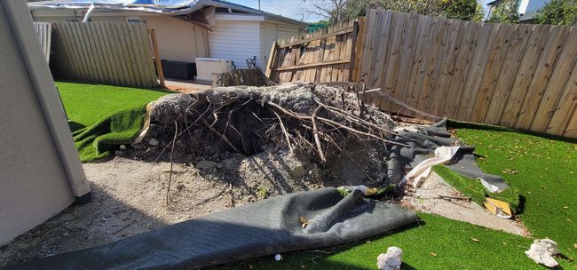 Debris pile in backyard next to artificial turf, with a damaged fence and house in the background.