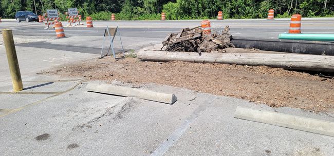 Construction site: dirt, exposed pipes, concrete bumpers, orange cones, vehicles on road.
