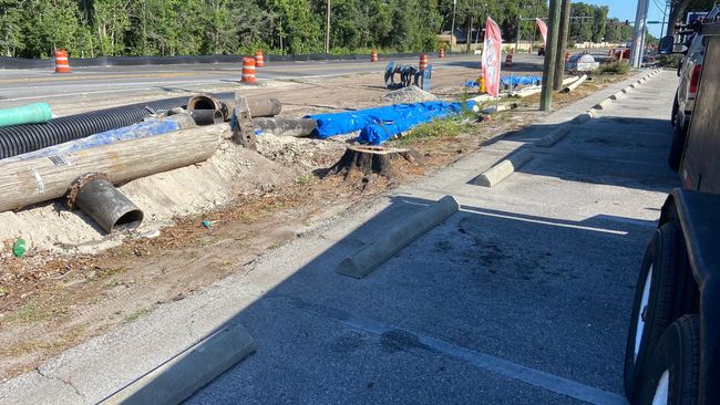 Construction site with exposed pipes, orange cones, and blue tarp; vehicle parked on the right.