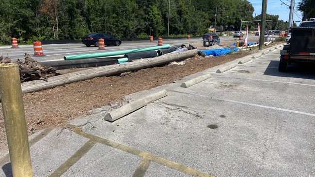 Road construction with open trench and pipes, parking area with barrier blocks and cones.