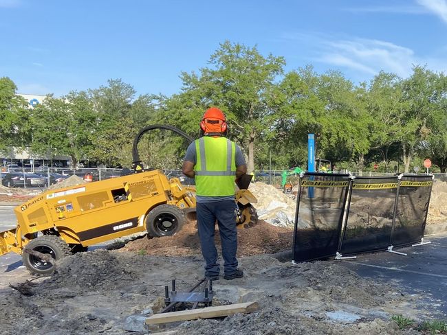 Man operating a yellow stump grinder at a construction site. He wears a hard hat and safety vest.