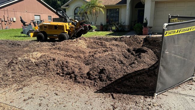 A stump grinder removing a tree stump in front of a house, surrounded by wood chips.