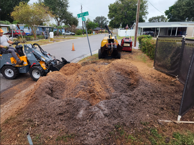 Tree removal crew: loader, chipper, and a large pile of mulch on a roadside.