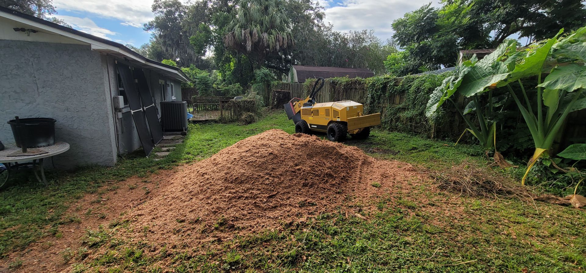 A yellow stump grinder is moving a pile of wood chips in a backyard.