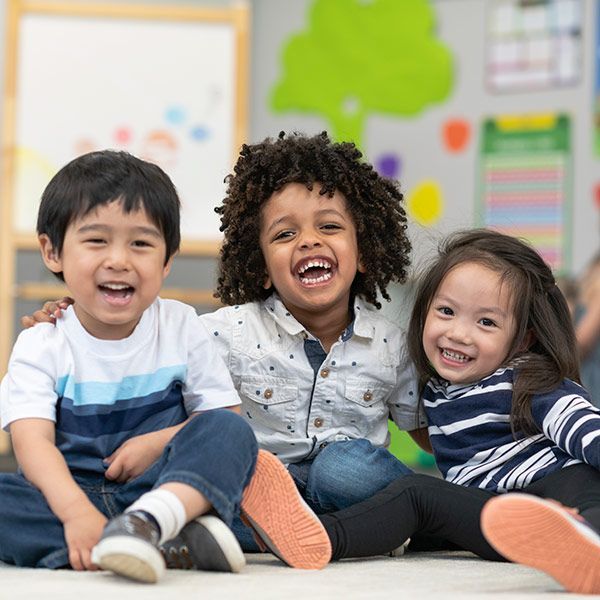 Three smiling children sitting on the floor in a bright classroom, laughing together