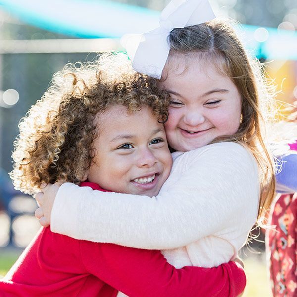 Two children hugging tightly and smiling outdoors, one in red and one in white.
