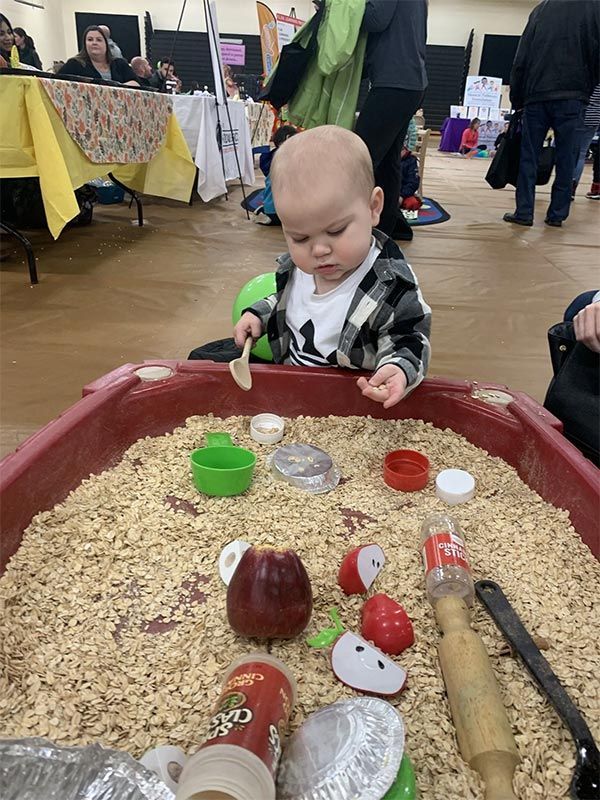 Toddler playing in a sensory bin with beans and toy utensils at an indoor event