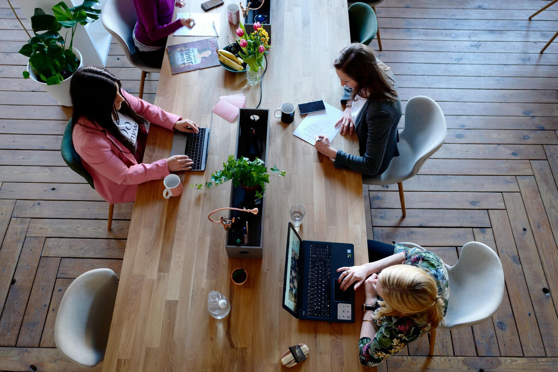 Four people working at a long wooden table, each with a laptop or papers.