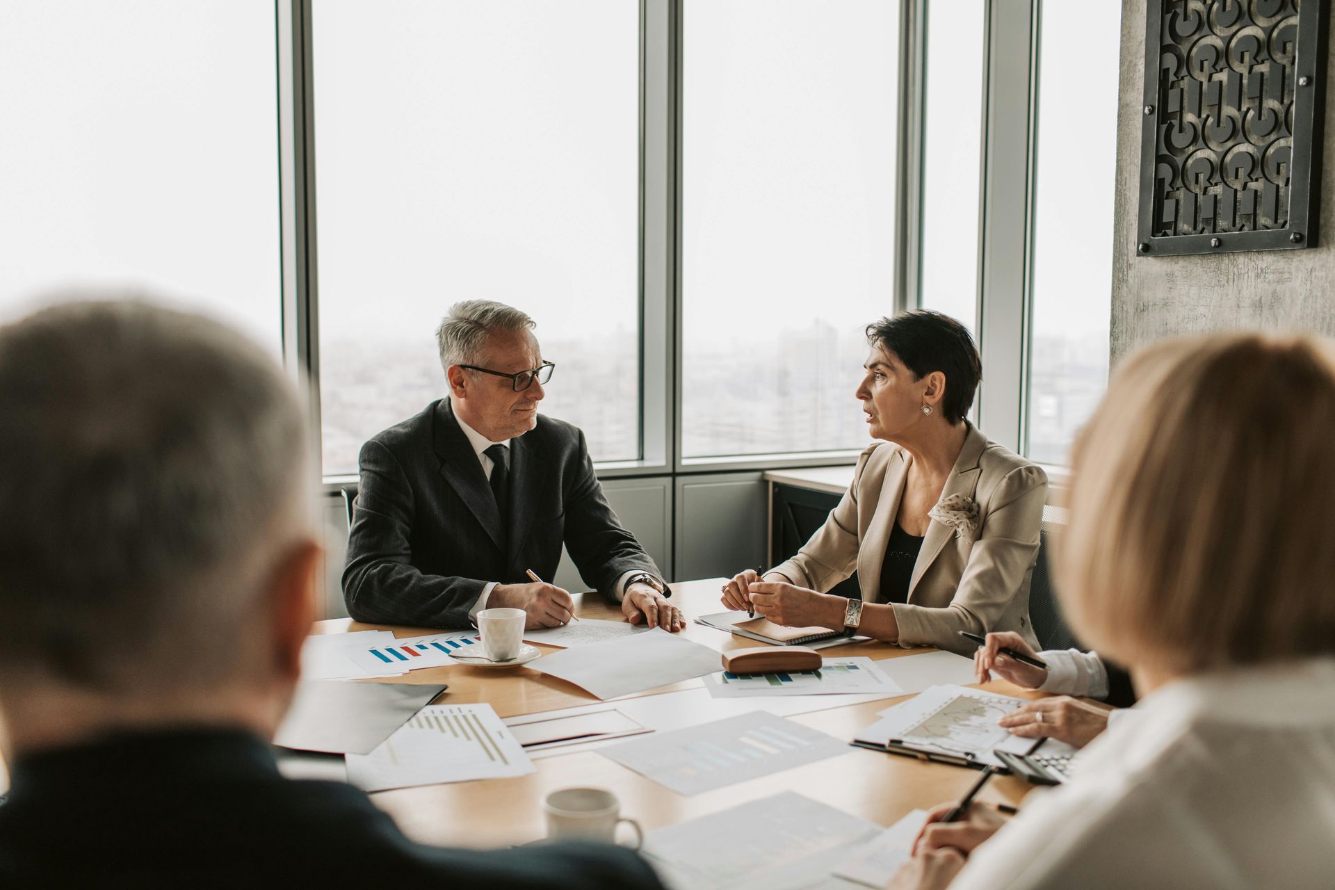 Business meeting in a modern office; people seated around a table, documents and coffee cups present, discussing a plan.