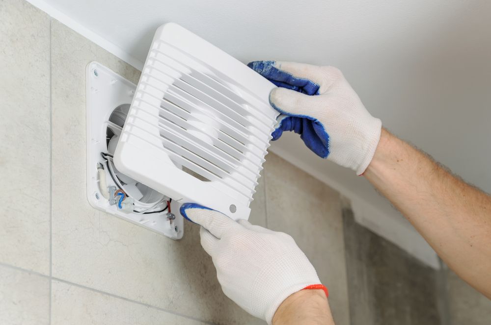Person Installing a White Bathroom Exhaust Fan on a Tiled Wall — Mazlin Electrical Services Pty Ltd in Kirwan, QLD