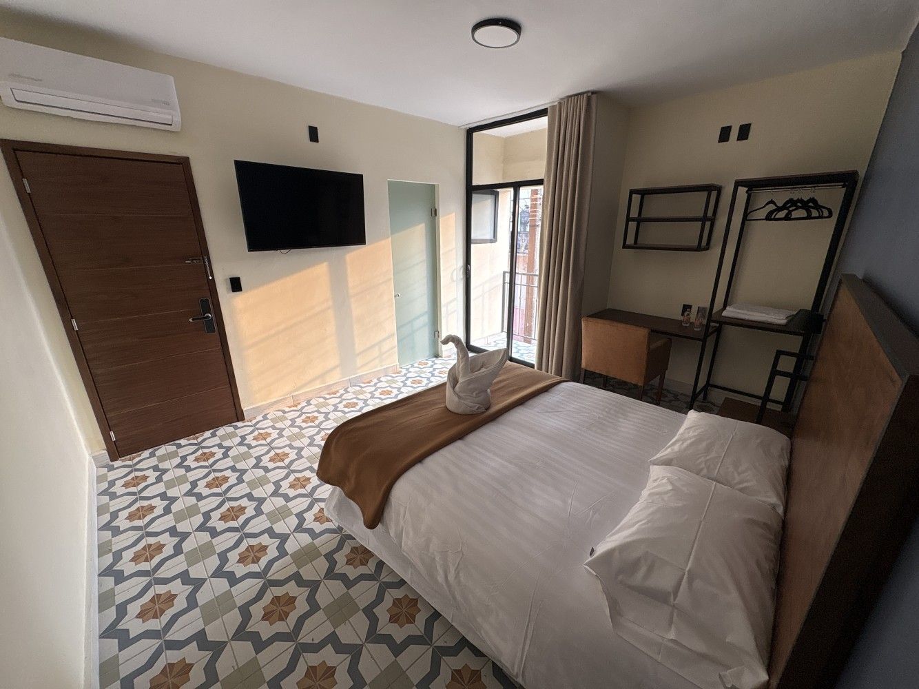 Dormitorio moderno con cama, televisor montado en la pared, suelo estampado y puerta abierta al balcón.